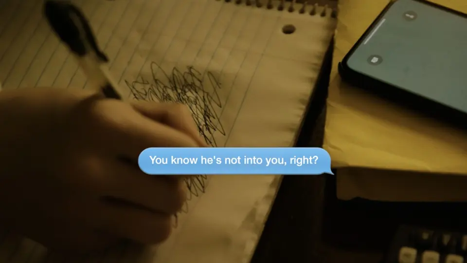 A hand writing in a notebook beside a phone showing a text message.