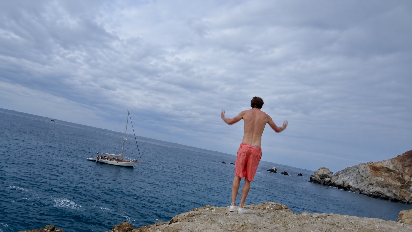 A man in red shorts standing on a rock, facing the sea.